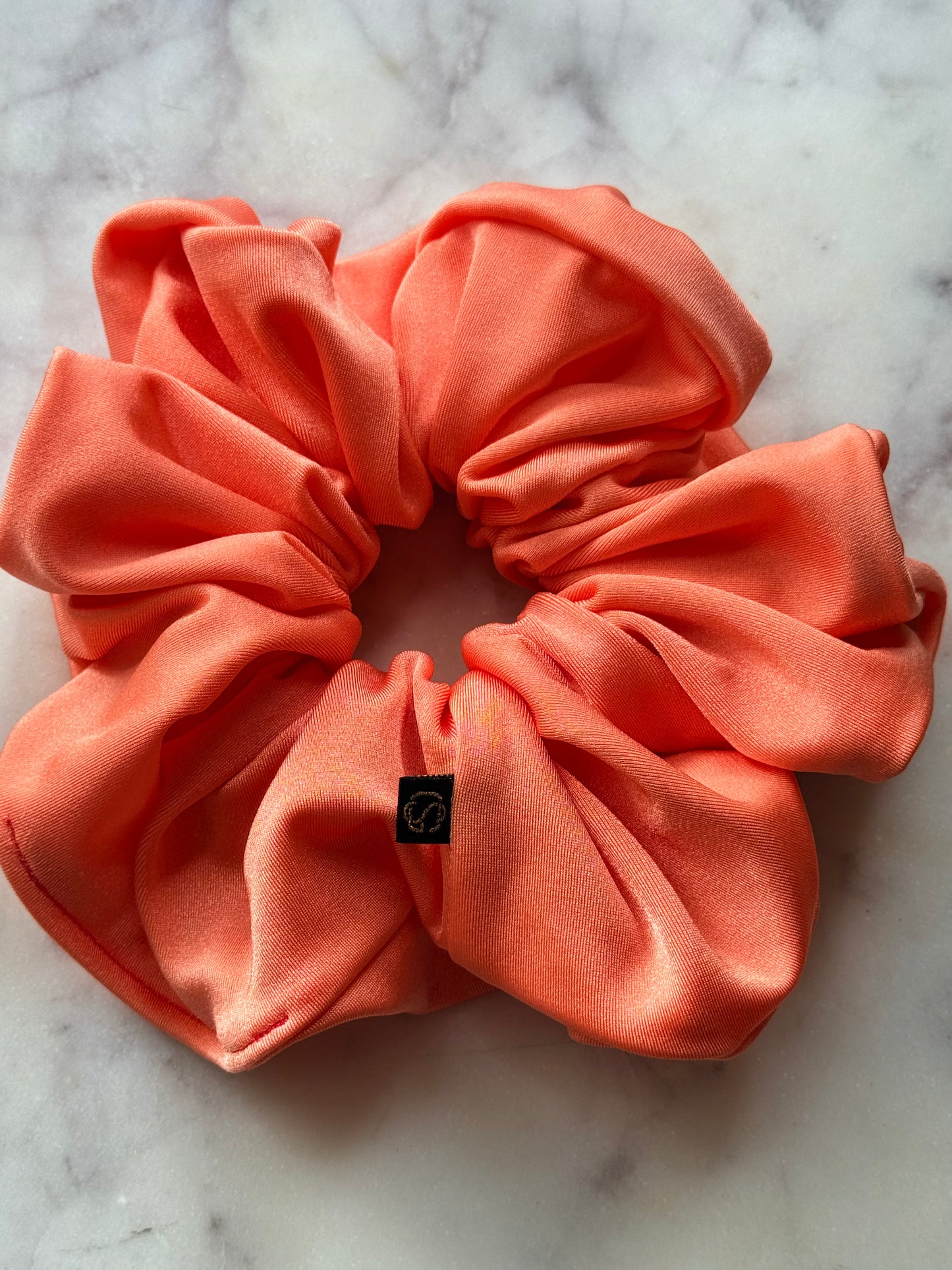 A coral pink satin scrunchie with a small black square logo tag, displayed on a white marble surface.