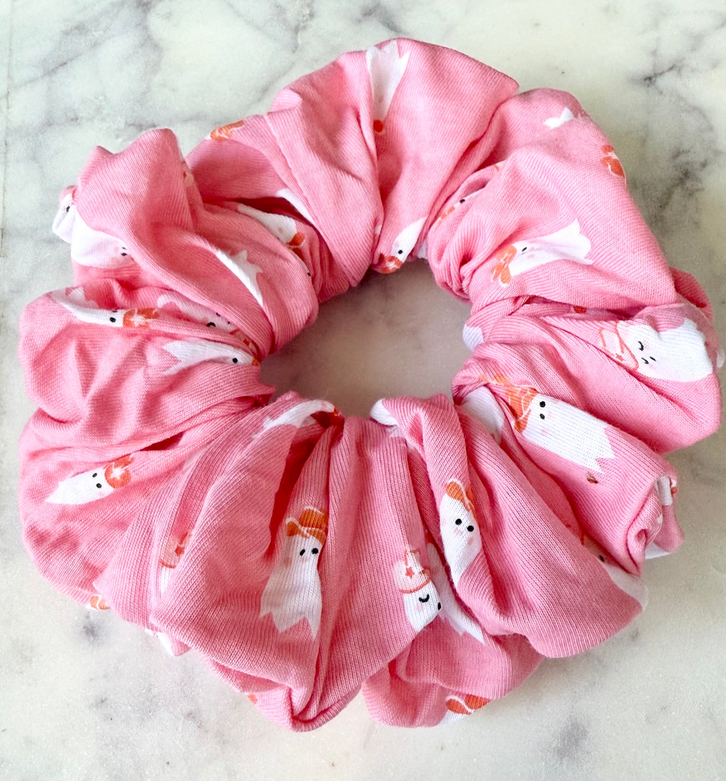 A pink fabric hair scrunchie with a white and orange cartoon ghost pattern, photographed flat on a light marble surface.