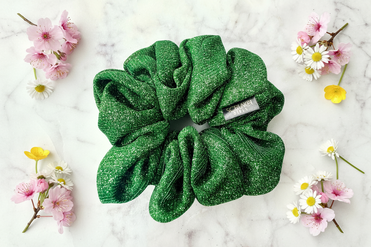 A green glittery scrunchie with a small white tag reading 'CHANEL PARIS' is arranged on a white marble surface with pink, white, and yellow flowers around it.