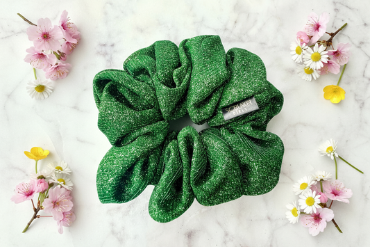 A green glittery scrunchie with a small white tag reading 'CHANEL PARIS' is arranged on a white marble surface with pink, white, and yellow flowers around it.