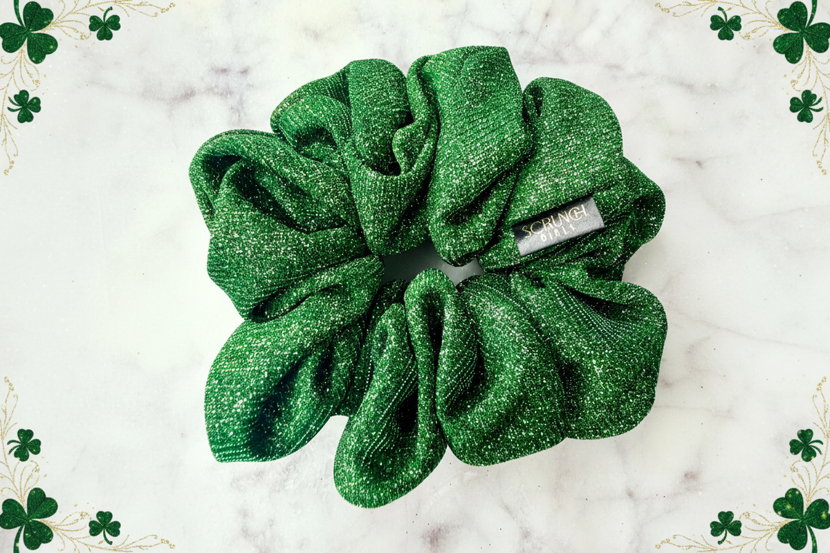 Green glittery scrunchie with a brand label on a light gray background with shamrock decorations.