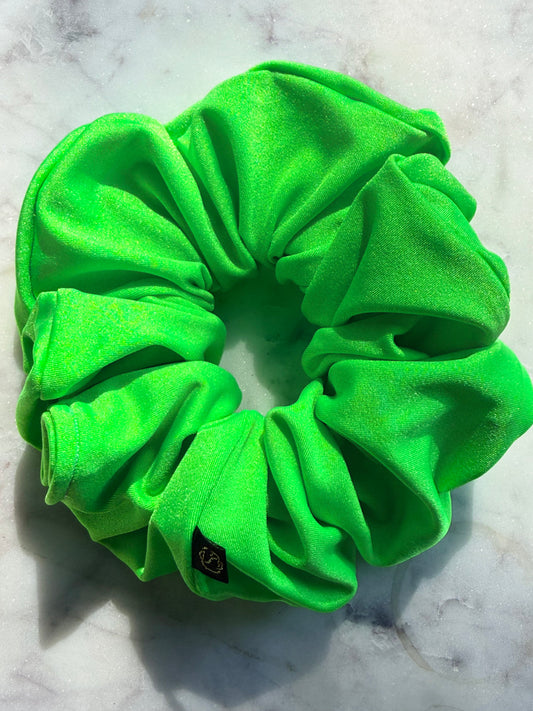 A bright neon green fabric scrunchie with a small black brand tag, resting on a light gray marble surface.