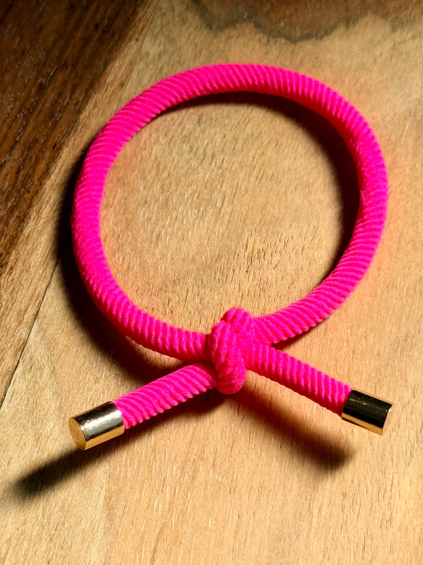 A bright pink braided cord bracelet with gold-toned metal end caps, arranged in a loop on a wooden surface.