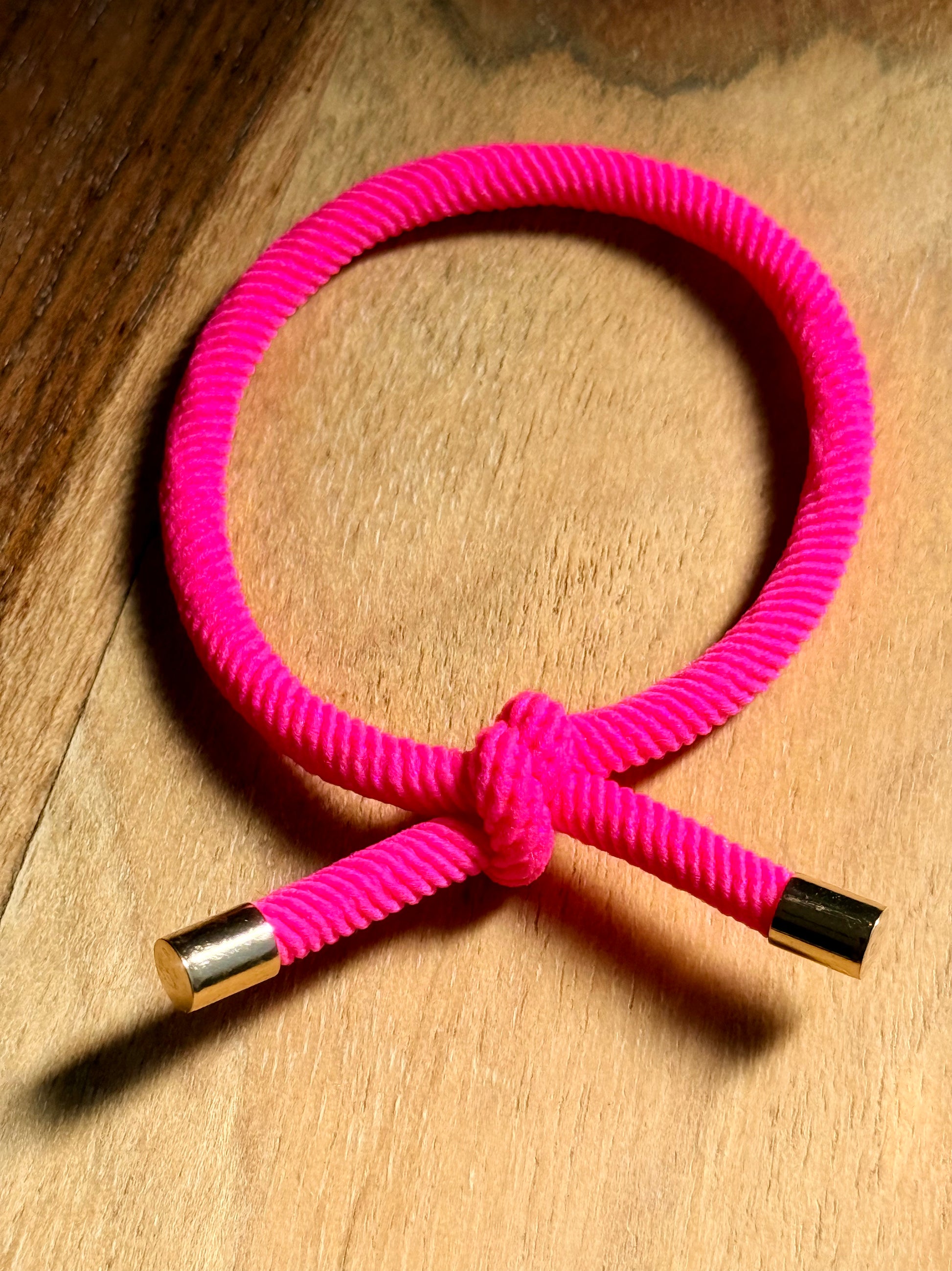 A bright pink braided cord bracelet with gold-toned metal end caps, arranged in a loop on a wooden surface.