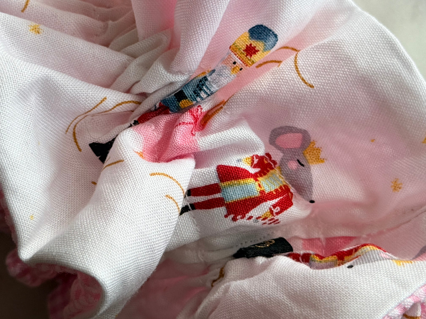 A close-up of a white fabric accessory, likely a baby bonnet or headband, featuring a pink ruffled trim and a whimsical printed pattern of cartoon characters in red, blue, yellow, and black.