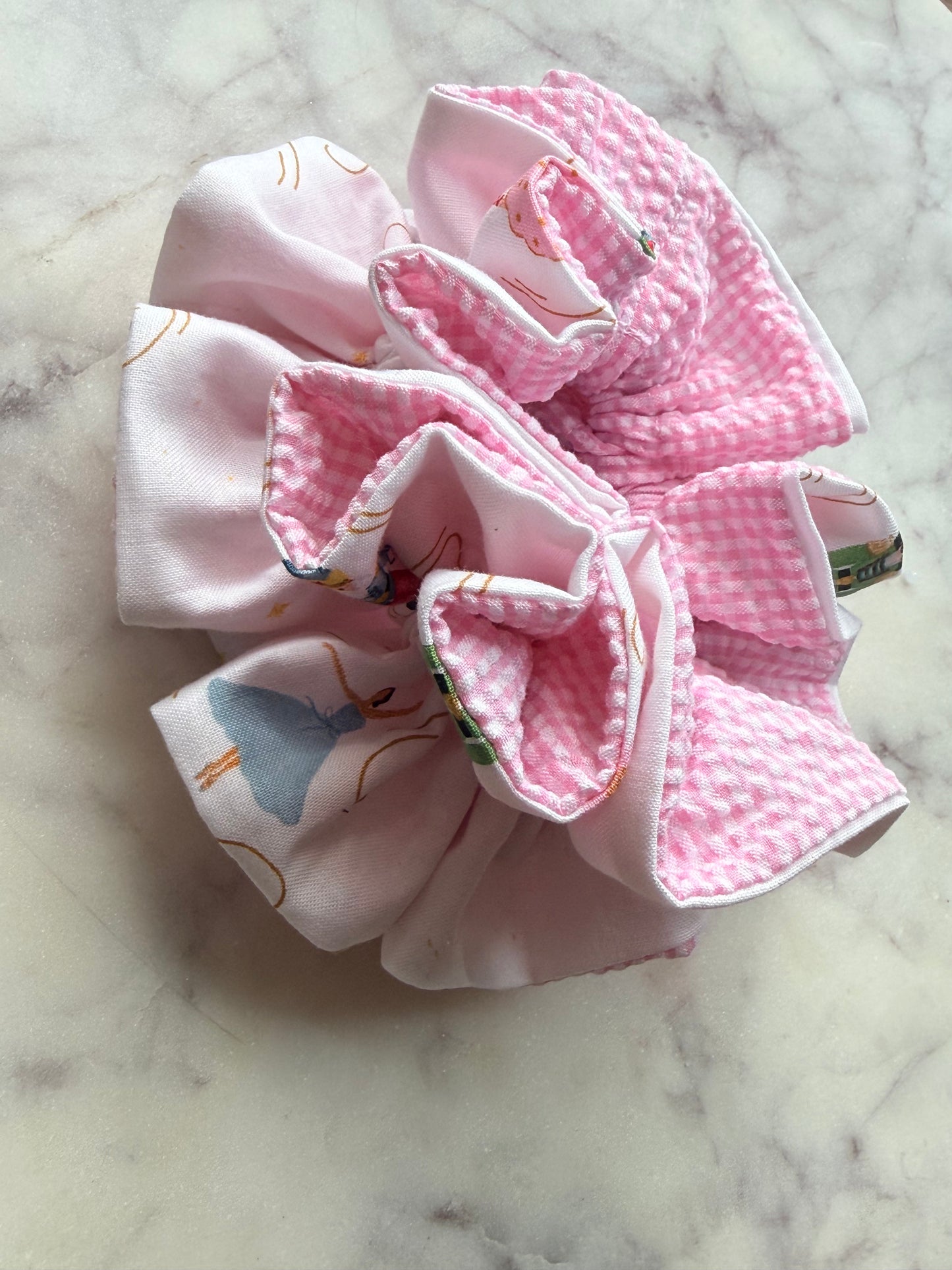 A pink fabric hair scrunchie with a gingham pattern and printed ballet dancer motifs, displayed on a light marble surface.