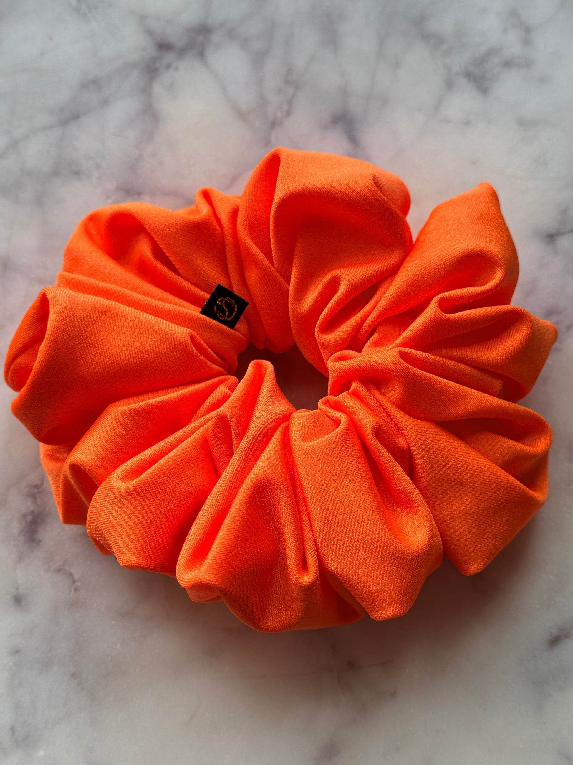 A bright orange fabric scrunchie with a small black brand tag, resting on a white and gray marble surface.